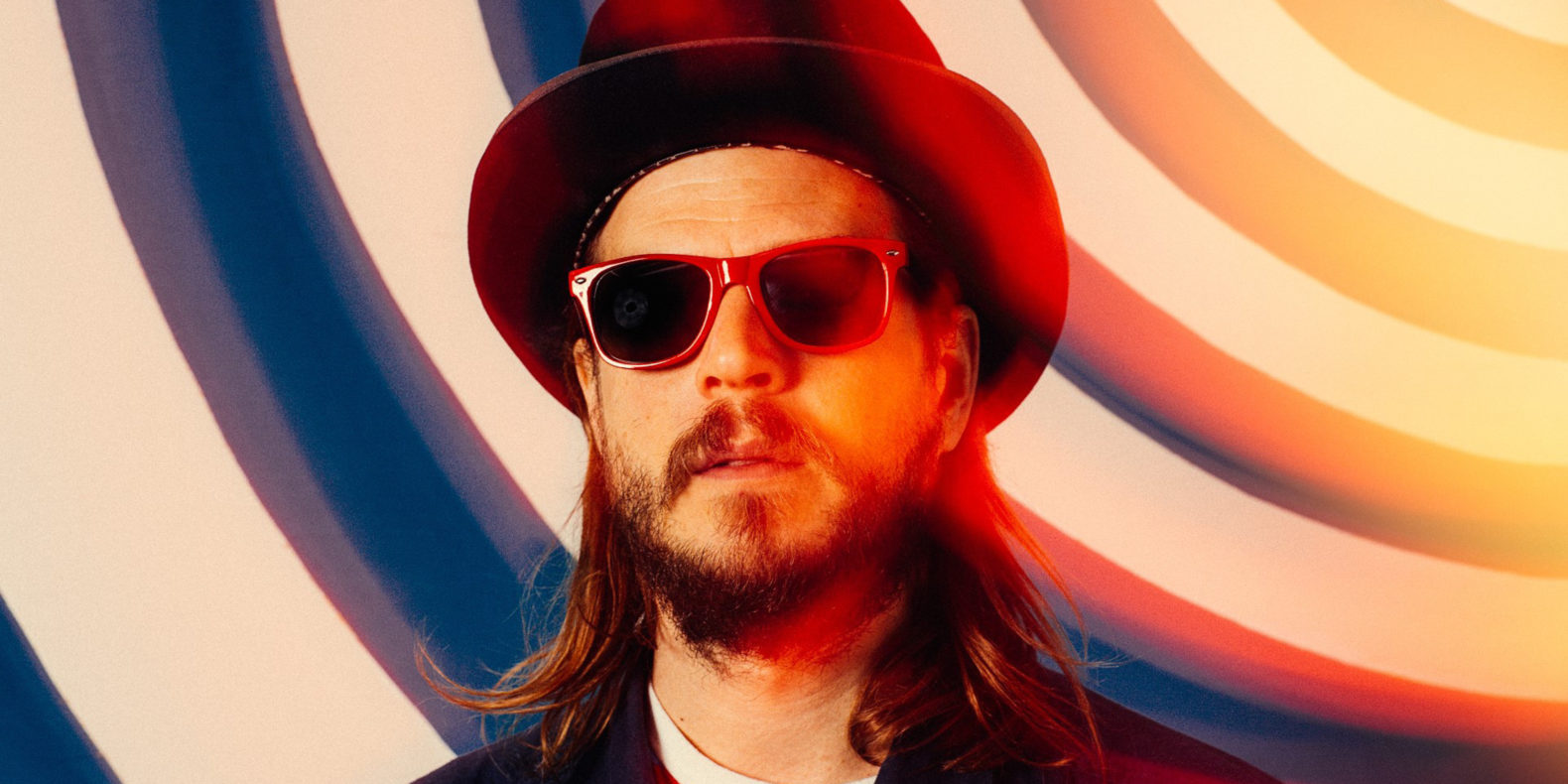 Piano player Marco Benevento in a top hat and sunglasses in front of a wall with curved blue and white stripes