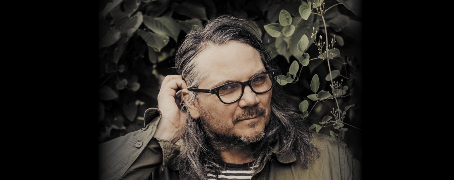 Jeff Tweedy benefit concert feature image, Jeff Tweedy in green shirt in front of green foliage tucking hair behind ear