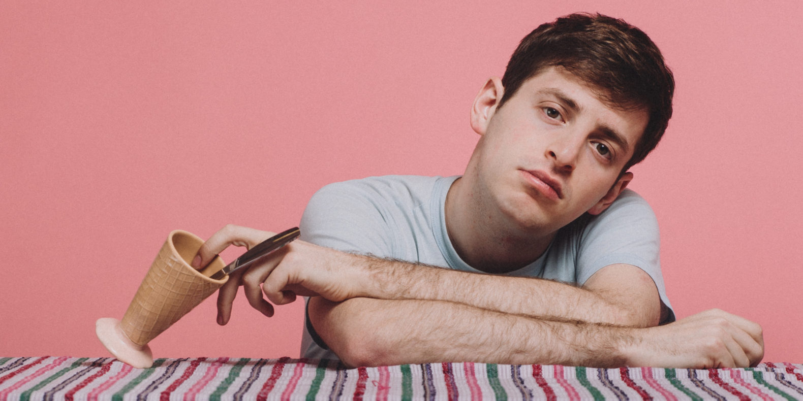 Alex Edelman feature image, Alex leaning on table with empty ice cream cup in front of pink background
