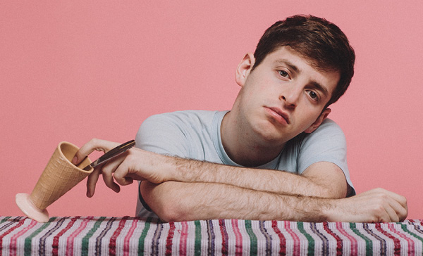 Alex Edelman feature image, Alex leaning on table with empty ice cream cup in front of pink background