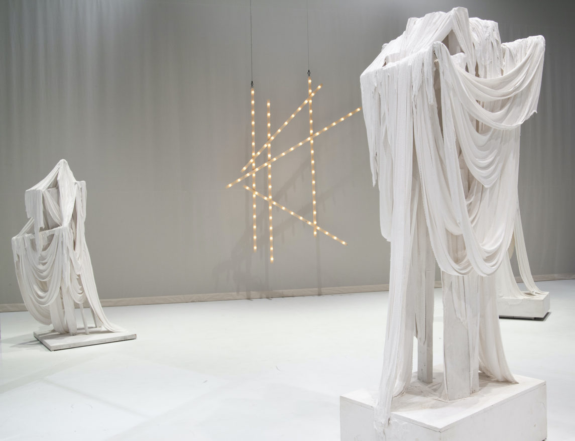 Marc Swanson feature image, sculptures that appear to be strips of draped white fabric with against white floor and wall, with LED lights on wall