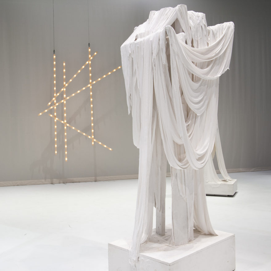 Marc Swanson feature image, sculptures that appear to be strips of draped white fabric with against white floor and wall, with LED lights on wall