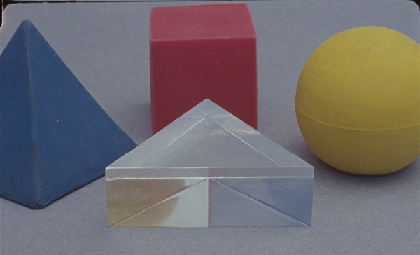 Yto Barrada feature image, Tree Identification for Beginners ( film still), picturing blue pyramid, red cube, yellow sphere, and clear triangle next to each other