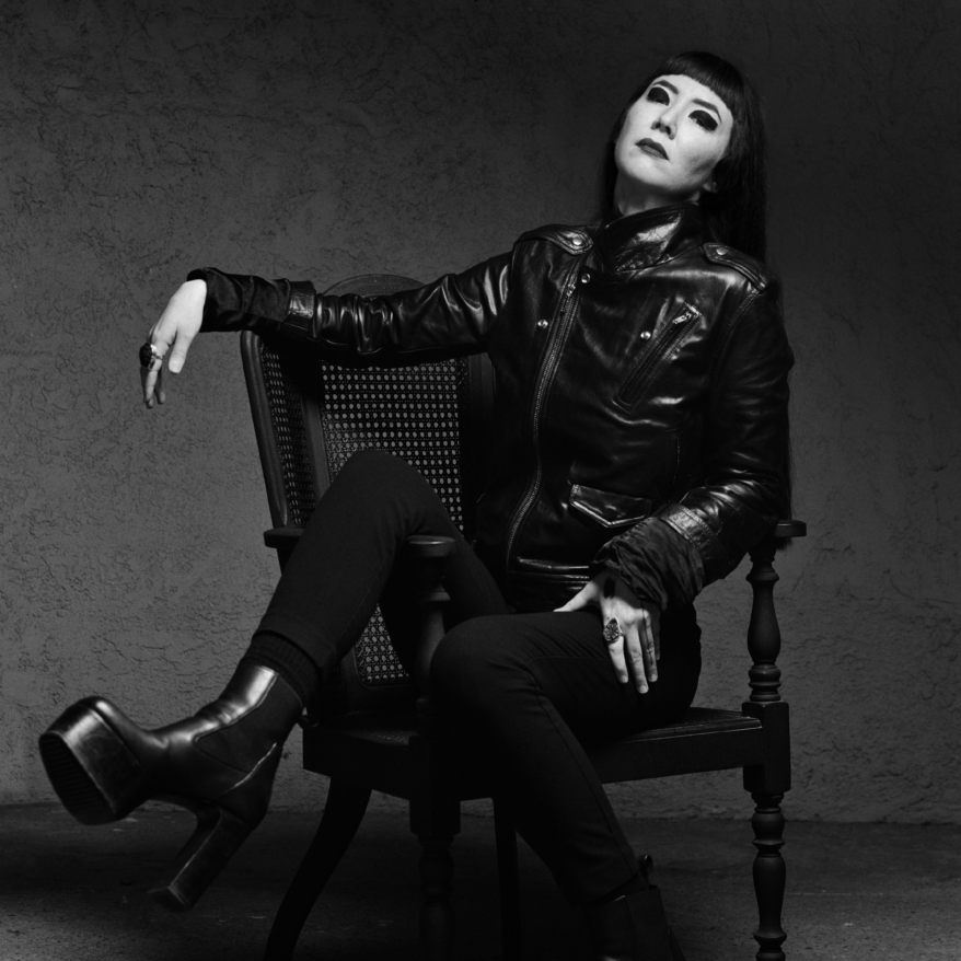 Johanna Hedva, dressed in all black leather, sits in a chair with their legs over the side, posed looking up at the sky.